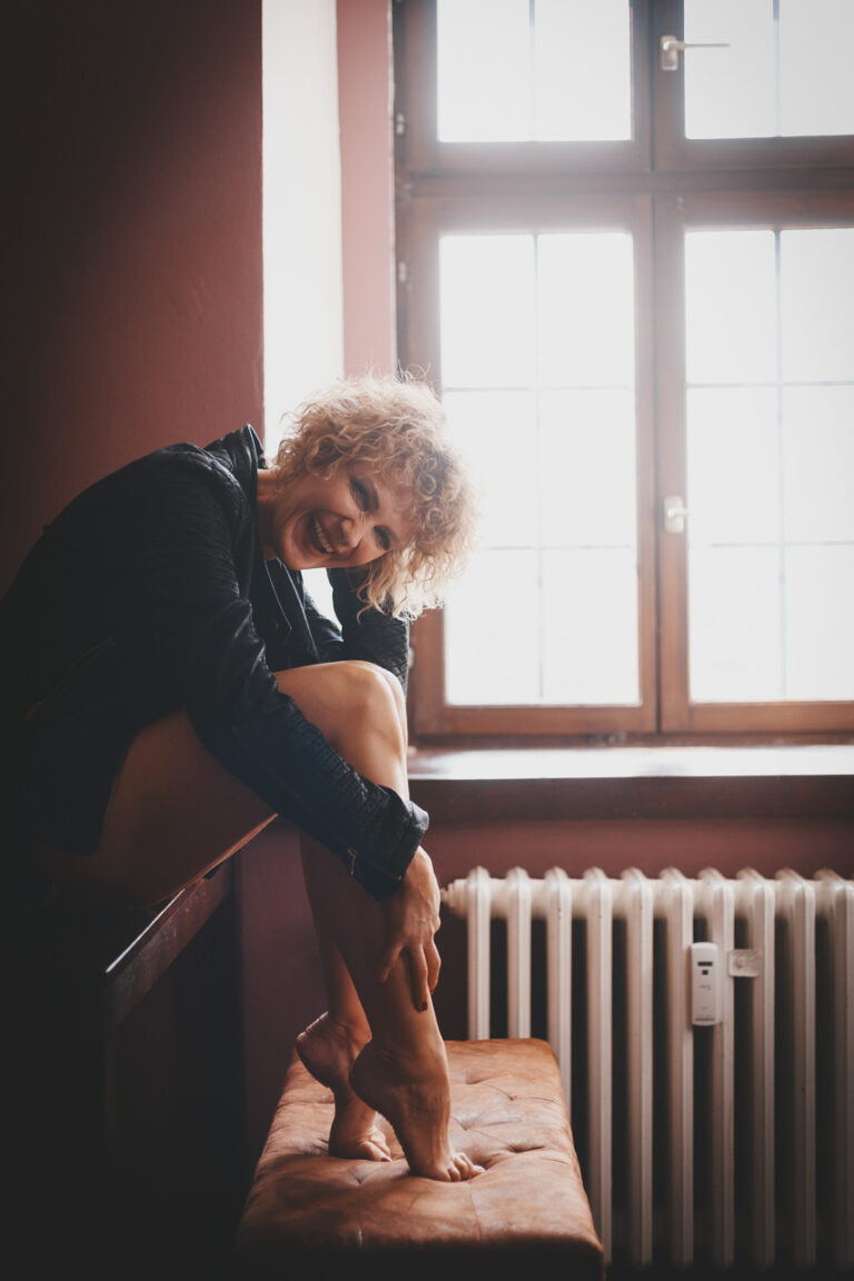 Frau mit Locken sitzt lachend vor einem hellen Fenster