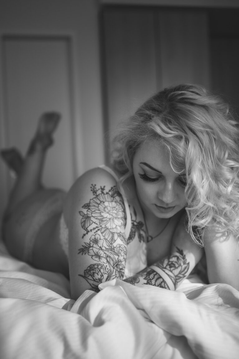 Frau mit Locken und Tattoos liegt auf weißer Bettwäsche, Schwarzweiß